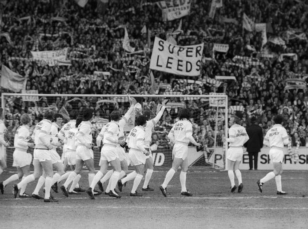 Historic Leeds United players walking onto the pitch with supporters in the stand.