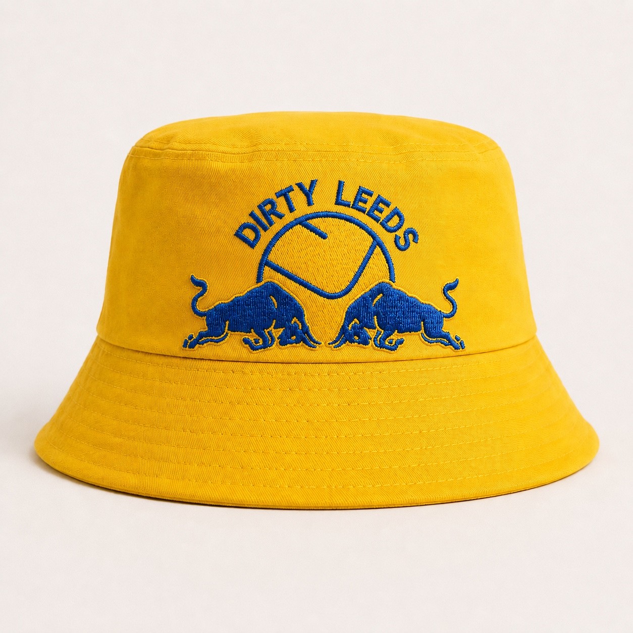 Bright yellow Dirty Leeds® bucket hat with blue embroidered Dirty Leeds text, smiley badge and Red Bull emblem on the front.